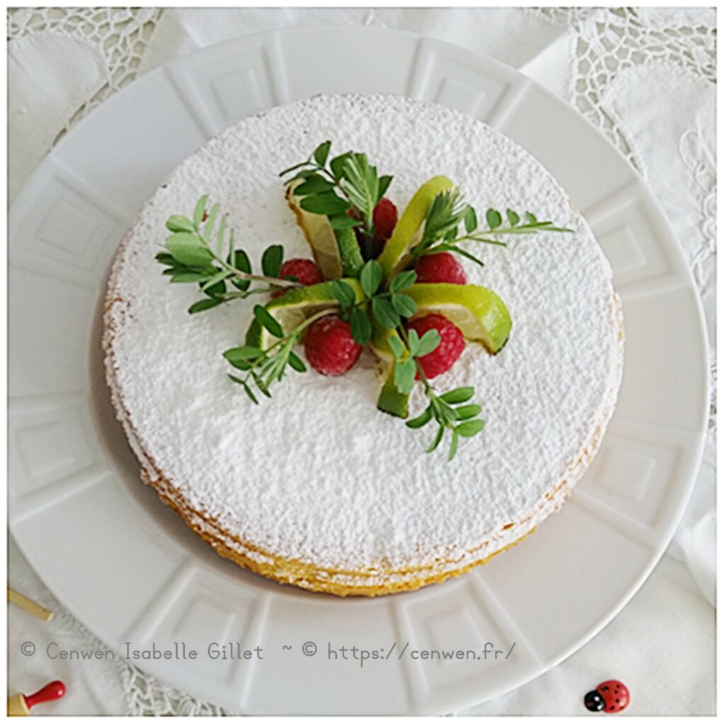 Gâteau rond saupoudré de sucre glace, décoré au centre de framboises fraîches, de rondelles de citron vert et de quelques feuilles.