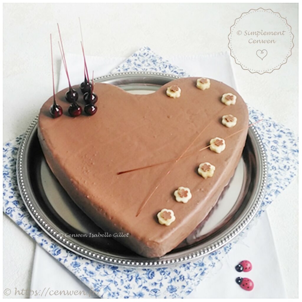 Gâteau en forme de cœur recouvert d’un glaçage chocolat au lait, décoré de fleurs découpées dans des rondelles de banane et de noisettes caramélisées.