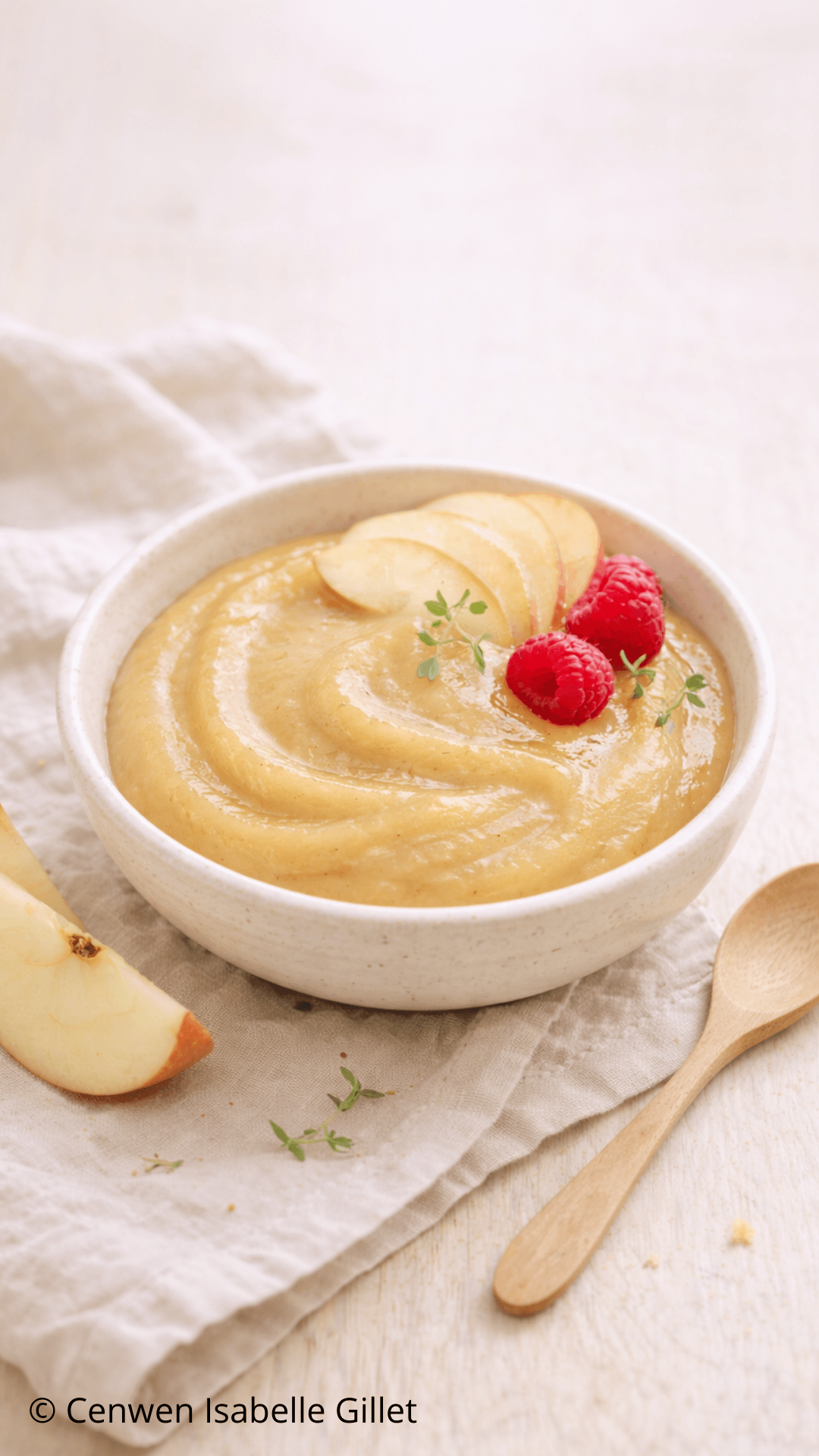 Bol de beurre de pommes lisse et doré, décoré de fines tranches de pomme, framboises fraîches et herbes, présenté sur un linge clair avec une cuillère en bois