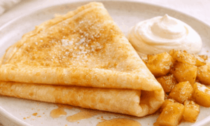 Crêpes pliées nappées de sirop, accompagnées de pommes caramélisées et d’une touche de crème fouettée.