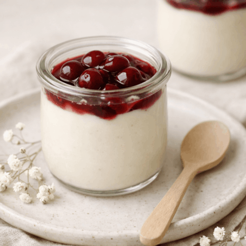 Petits pots de crème au lait d'amande nappés de cerises amarena au sirop, présentés dans un pot en verre sur une assiette en céramique blanche avec une cuillère en bois.